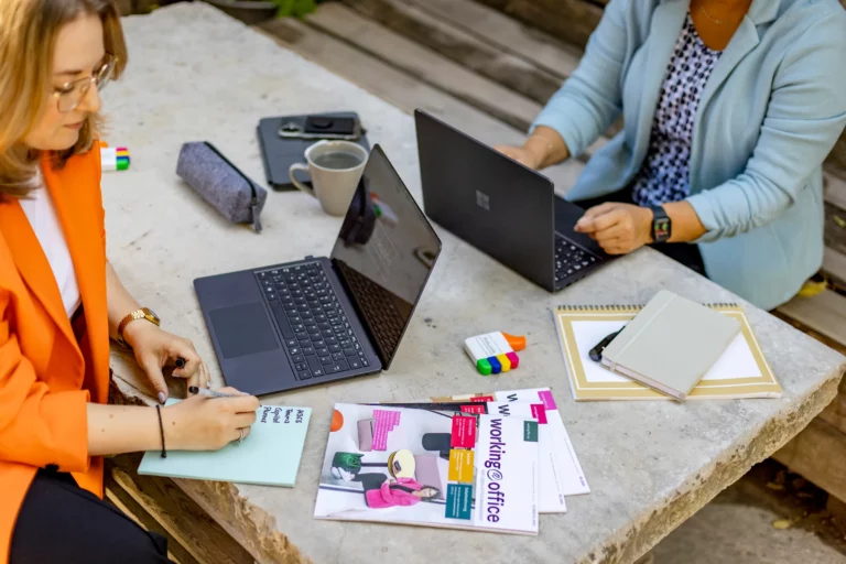 Katja Beckmeyer und Dominique Woith arbeiten mit Laptops und Unterlagen an einem Tisch im Freien
